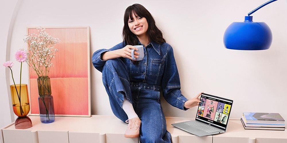 Kobieta w jeansowym kombinezonie siedzi na szafce z filiżanką kawy, obok laptop ASUS Vivobook S14 z wyświetloną galerią graficzną, po lewej wazon z kwiatami i obraz, po prawej niebieska lampa i stos książek, jasne, kreatywne wnętrze. - Opis serii VivoBook 