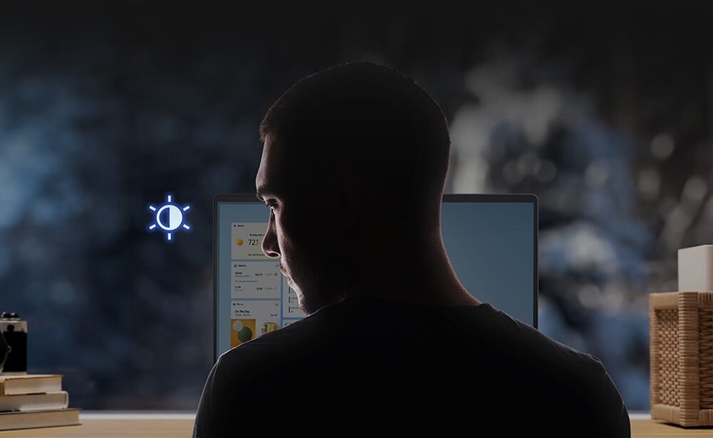 Osoba siedzi przed laptopem z widocznym ekranem, na którym wyświetlane są różne aplikacje. Obok ekranu znajduje się symbol jasności w postaci słońca z promieniami.