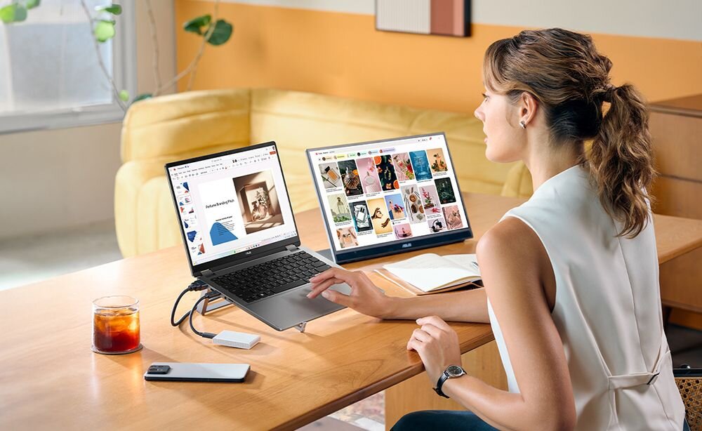 Osoba siedzi przy biurku z otwartym laptopem i dodatkowym monitorem, na którym wyświetlane są obrazy. Na biurku znajduje się również szklanka z napojem, smartfon i notes.