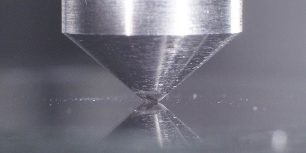 Zbliżenie na metalowy stożek dotykający gładkiej powierzchni, z widocznym odbiciem na tej powierzchni.