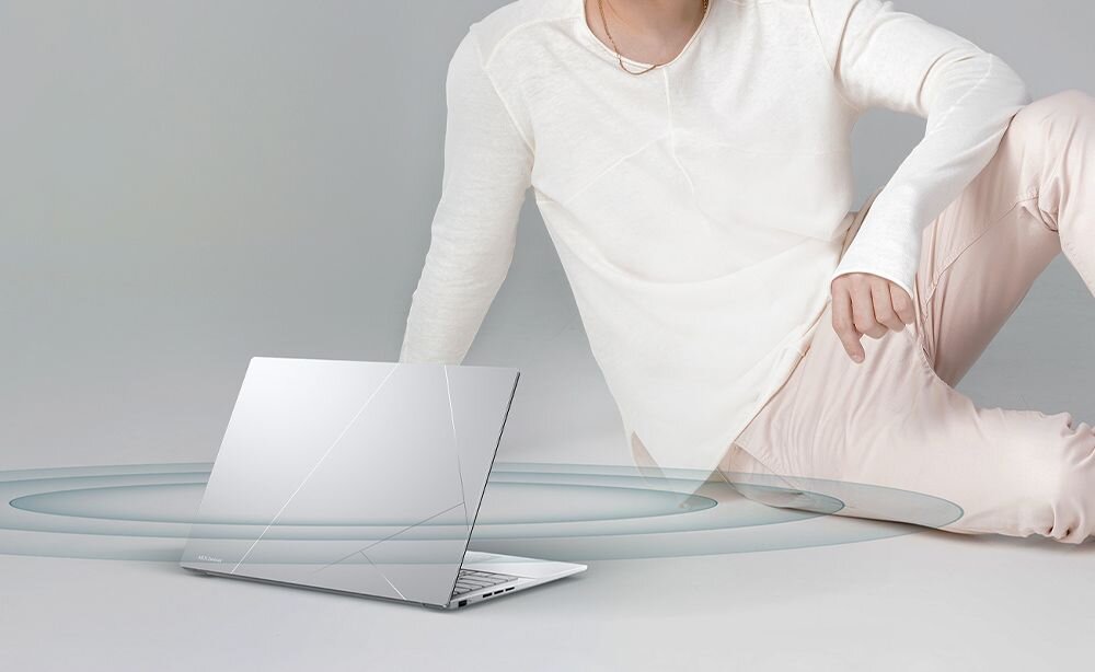 Na obrazie widać otwarty laptop o srebrnej obudowie z delikatnymi liniami na pokrywie. Obok laptopa siedzi osoba ubrana w jasne ubrania, a wokół laptopa widoczne są stylizowane fale dźwiękowe.