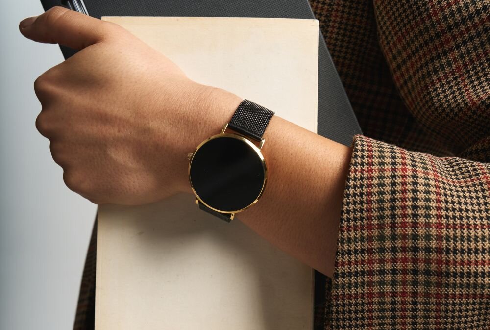 Osoba trzyma książkę, na nadgarstku ma czarno-złoty smartwatch z okrągłą tarczą i metalową bransoletą. Ubrana jest w marynarkę w kratę.