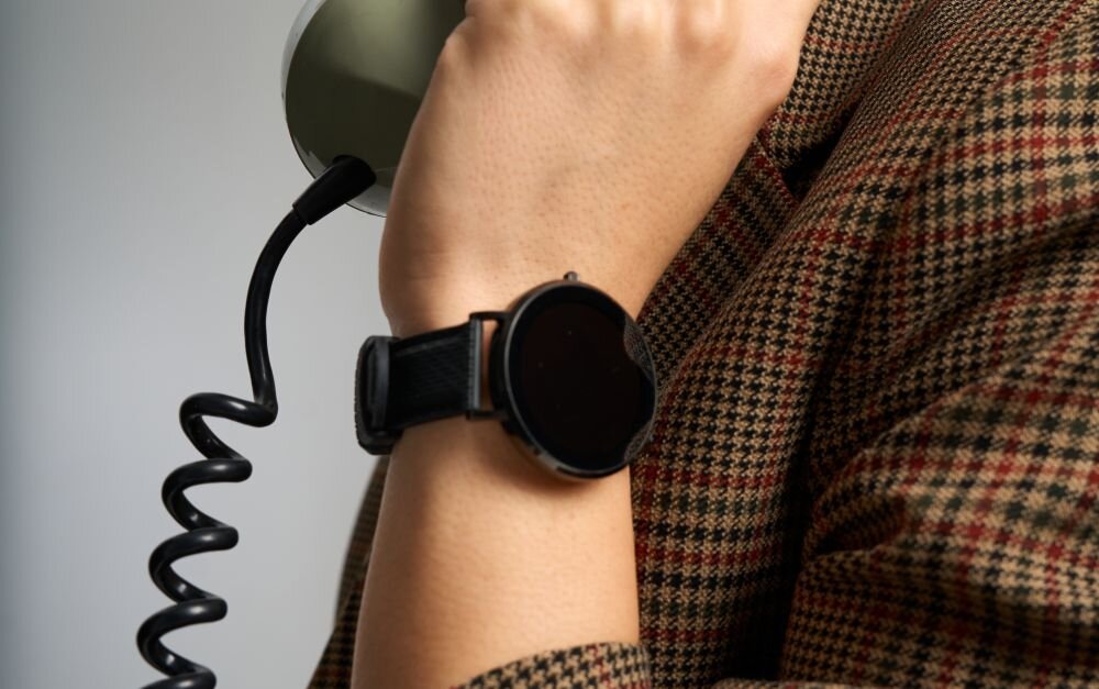 Osoba trzymająca telefon przy uchu, na nadgarstku ma czarno-złoty smartwatch z okrągłą tarczą. Ubrana jest w marynarkę w kratę.