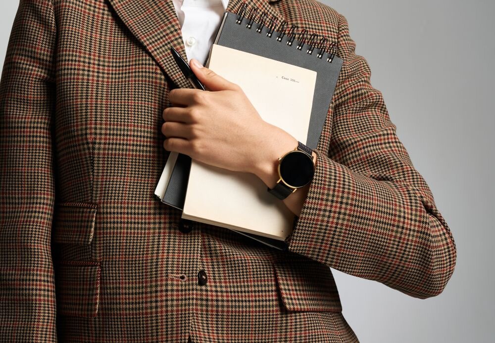 Osoba w kraciastej marynarce trzyma notes i długopis, na nadgarstku ma czarno-złoty smartwatch. Notes ma czarną okładkę i widoczny napis \'Color plan\'.
