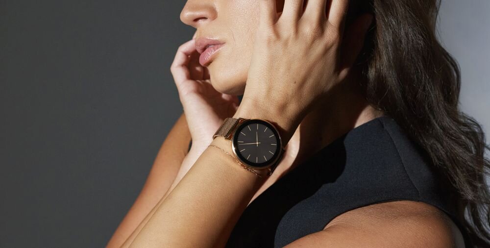 Osoba nosi złoty smartwatch z czarną tarczą i minimalistycznym designem. Tło jest neutralne, a osoba ma ciemne włosy i ciemny strój.