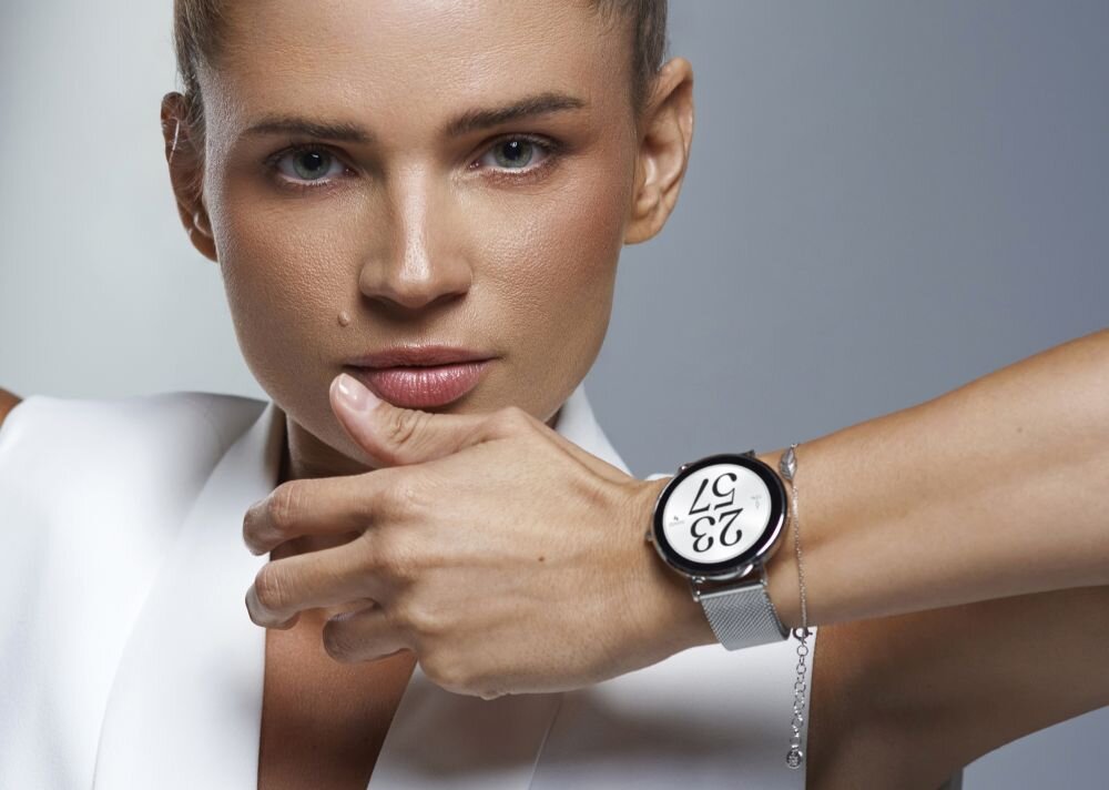 Osoba prezentuje smartwatch na nadgarstku, z wyraźnym wyświetlaczem pokazującym cyfry 23 i 57. Zegarek ma metalową bransoletę i okrągłą tarczę.