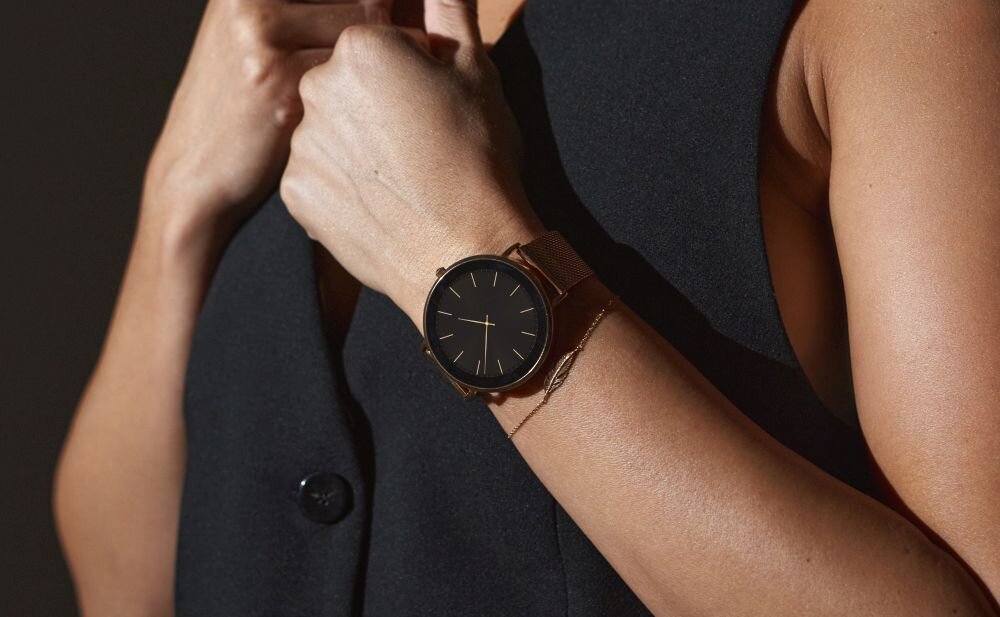 Osoba nosi złoty smartwatch z czarną tarczą i minimalistycznym designem. Ubrana jest w ciemną odzież bez widocznych wzorów.