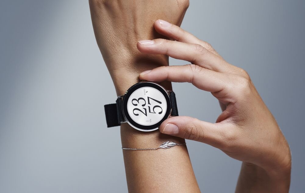 Osoba zakłada smartwatch z okrągłym wyświetlaczem, na którym widoczna jest godzina 23:57. Zegarek ma czarny pasek i srebrną kopertę.