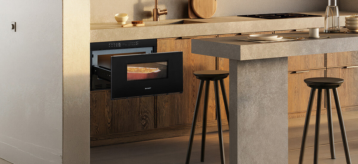 Kuchenka mikrofalowa SHARP RINDS333FB Przedstawiono kuchnię z wysuniętą szufladową kuchenką mikrofalową SHARP, w której podgrzewane jest danie w naczyniu żaroodpornym. Całość utrzymana jest w ciepłej, minimalistycznej aranżacji z drewnianą zabudową szybki start, stage cooking, wydłużenie czasu gotowania o 30 sekund, ustawienia wstępne, timer do 99:99, zegar, tryb oszczędzania energii, funkcja chłodzenia, czujnik wilgoci rozmrażanie wagowe