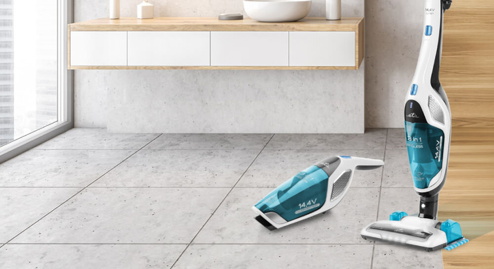 Na zdjęciu widać dwa odkurzacze w nowoczesnym wnętrzu z jasnymi płytkami podłogowymi i drewnianą szafką. Na odkurzaczach widoczny jest tekst \'14.4V\' oraz \'3 in 1\'.