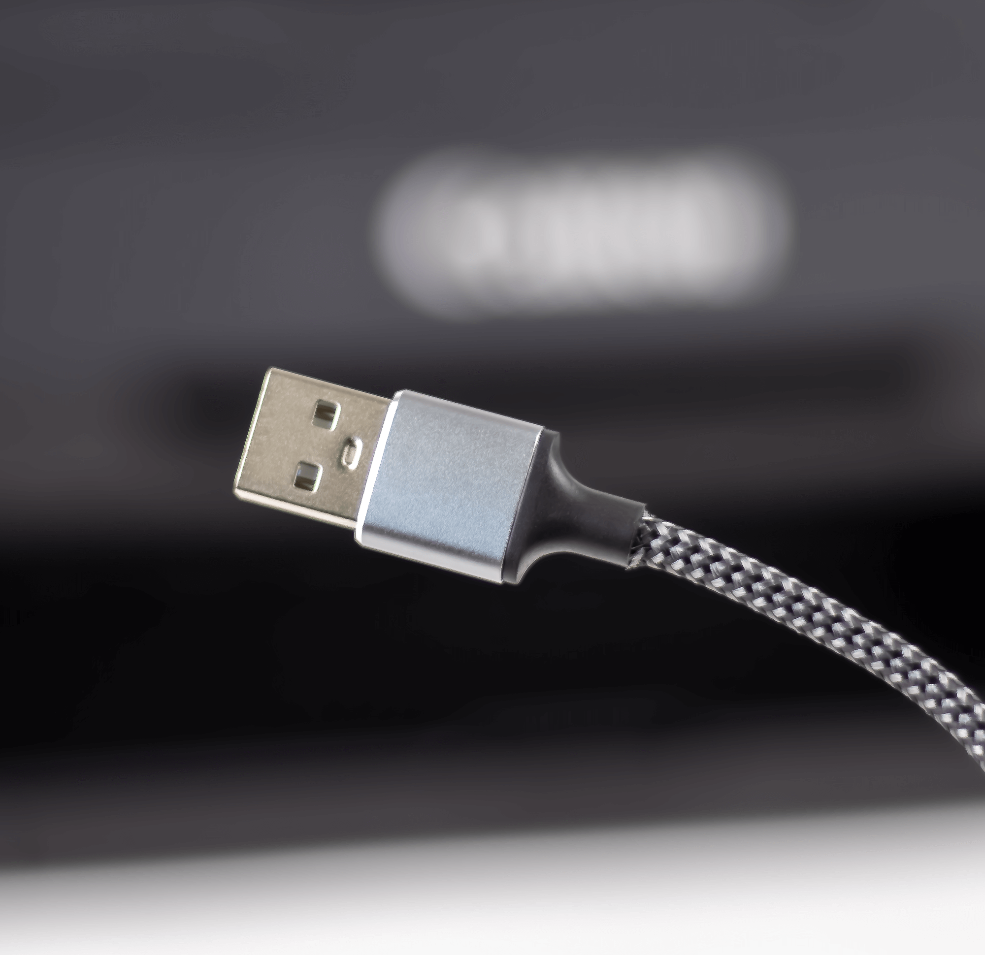 Zbliżenie na końcówkę kabla USB-A z metalową obudową i plecionym przewodem. W tle widoczne jest rozmyte logo producenta.