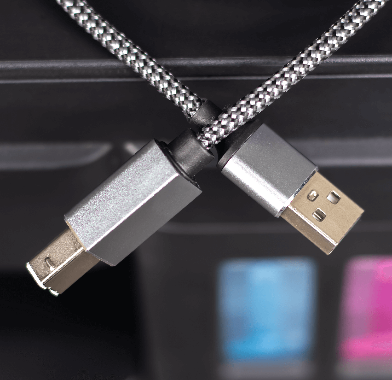 Na obrazie widoczny jest czarno-srebrny kabel USB z końcówkami USB-A i USB-B. Kabel ma plecioną osłonę i jest podłączony do urządzenia z kolorowymi elementami w tle.