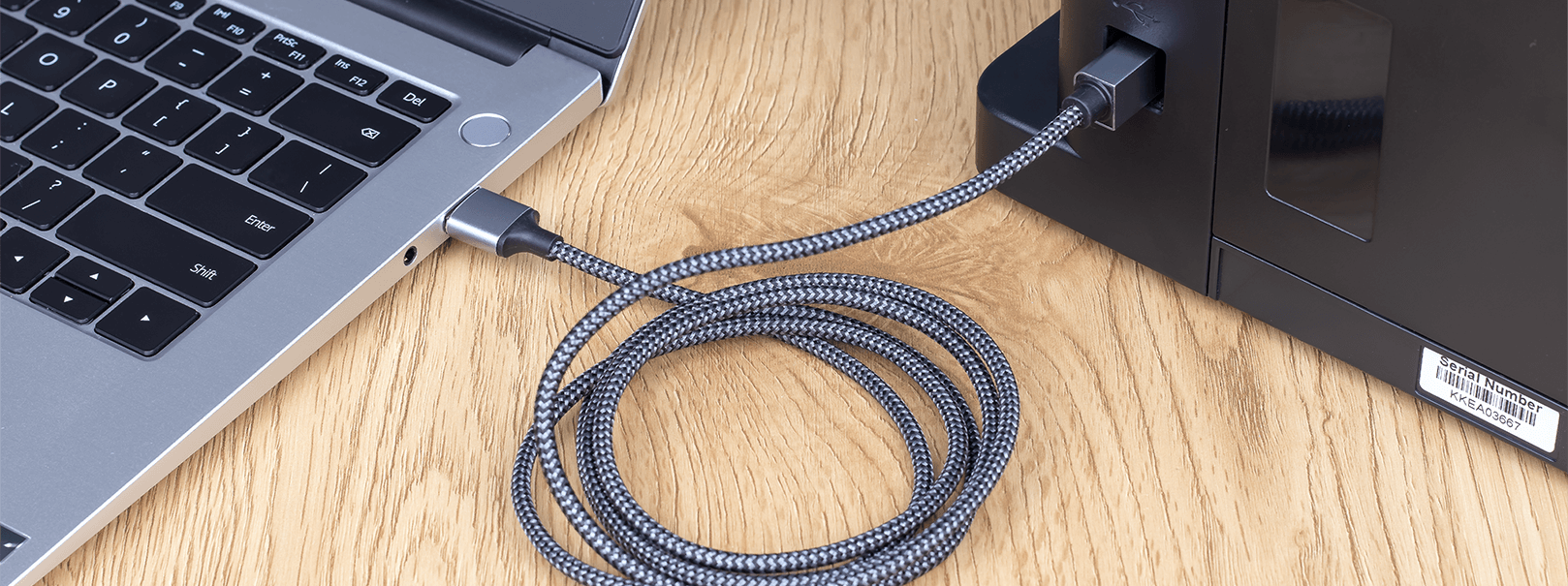 Na drewnianym blacie znajduje się laptop oraz urządzenie z podłączonym czarno-srebrnym kablem USB, który jest zwinięty w pętlę.