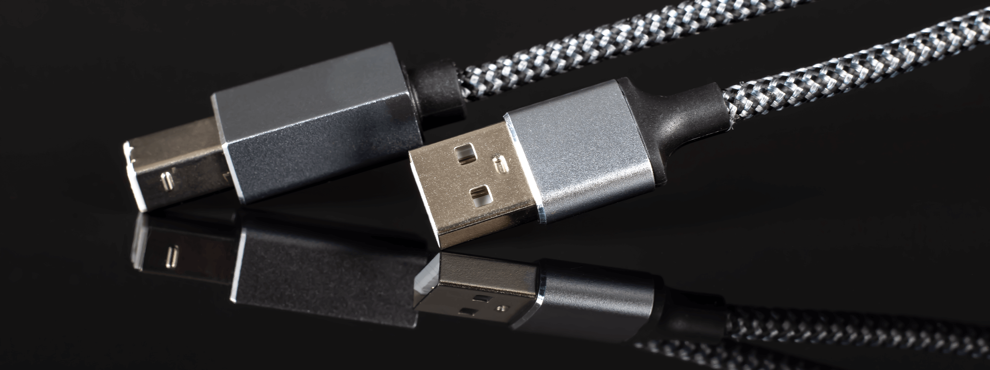 Na obrazie widoczne są dwa końce kabla USB z metalicznymi wtyczkami, jeden USB-A i drugi USB-B, z plecionym czarno-srebrnym przewodem. Tło jest czarne, a kabel odbija się na błyszczącej powierzchni.