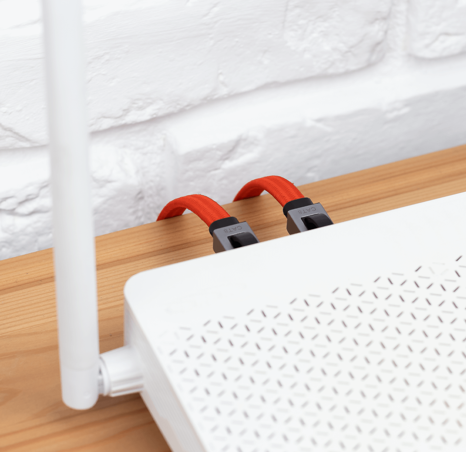 Na drewnianym blacie stoi biały router z widocznymi otworami wentylacyjnymi, do którego podłączone są dwa czerwone kable sieciowe z oznaczeniem \'CAT8\'.