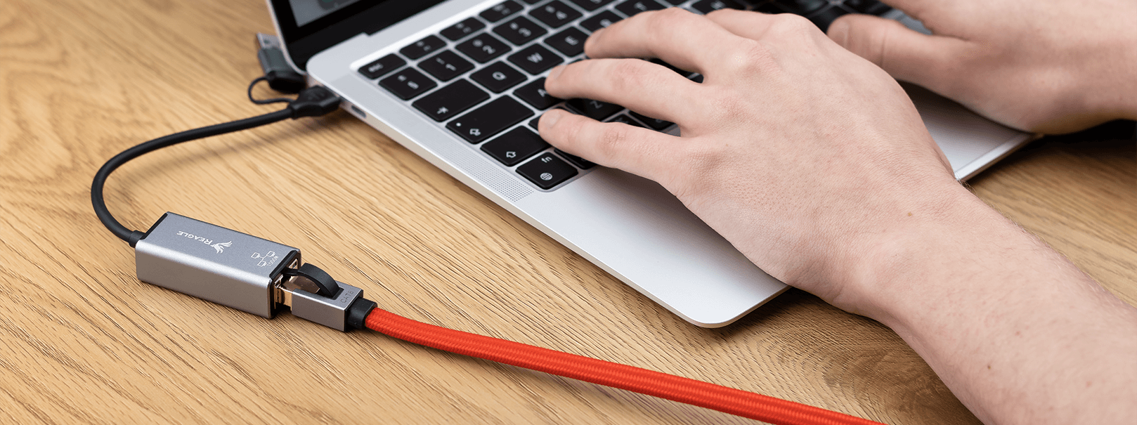 Osoba pisze na laptopie, do którego podłączony jest czerwony kabel sieciowy z metalową końcówką. Na końcówce widoczne jest logo producenta i napis \'40 Gb\'.