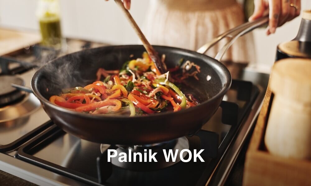 PŁYTA BEKO HILL75235S Na grafice widać dużą patelnię wok ustawioną na palniku gazowym, w której smażą się kolorowe warzywa, m.in. papryka i cebula. Na dole pojawił się biały napis „Palnik WOK”, podkreślający, że palnik jest szybko, gotowo smażenia palnik wok szybkie smażenie wysoka moc