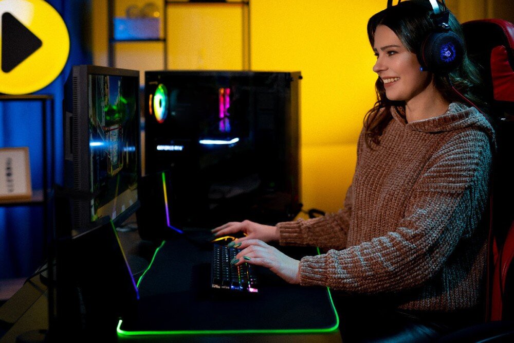 Osoba siedzi przy biurku, używając komputera z podświetlaną klawiaturą i słuchawkami na uszach. W tle widać obudowę komputera z podświetleniem RGB oraz żółtą ścianę z logo w kształcie strzałki.
