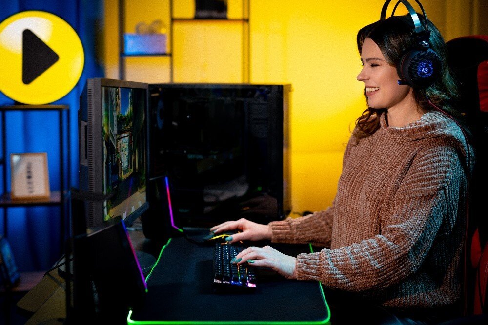 Komputer MAD DOG Powered by MSI F321R-I05DR32 Kobieta w słuchawkach grająca na komputerze stacjonarnym. Siedzi przy biurku z podświetlaną klawiaturą i monitorem, w tle niebiesko-żółte oświetlenie i regał, opis