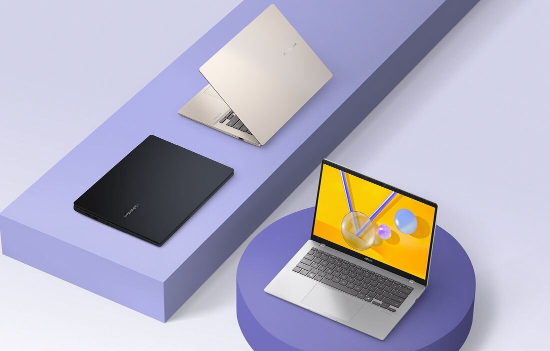Na obrazie widoczne są trzy laptopy w różnych kolorach: czarnym, złotym i srebrnym, ustawione na fioletowych platformach. Ekran jednego z laptopów wyświetla kolorową grafikę z geometrycznymi kształtami.