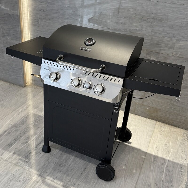 Czarny grill gazowy z trzema pokrętłami na panelu przednim i logo \'VELACO\' na pokrywie. Grill stoi na dwóch kołach, a po bokach znajdują się składane półki.