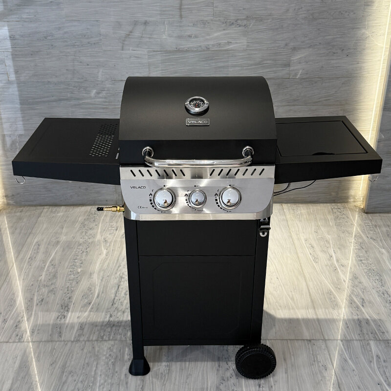 Czarny grill gazowy z trzema pokrętłami na srebrnym panelu, z logo \'VELACO\' na pokrywie. Grill stoi na marmurowej podłodze, a po bokach ma dwa składane blaty.