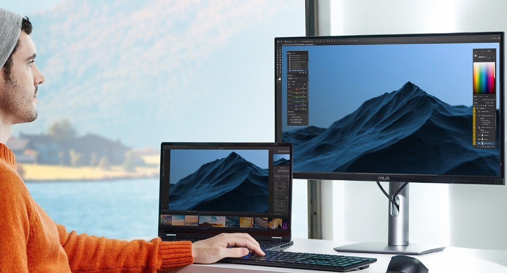 Monitor ASUS ProArt Display PA27JCV Osoba pracuje przy biurku z laptopem i podłączonym monitorem, na ekranach wyświetlony ten sam obraz górski w programie graficznym; za oknem widoczny krajobraz z wodą i wzgórzami, matryca, powłoka antyodblaskowa, widoczność