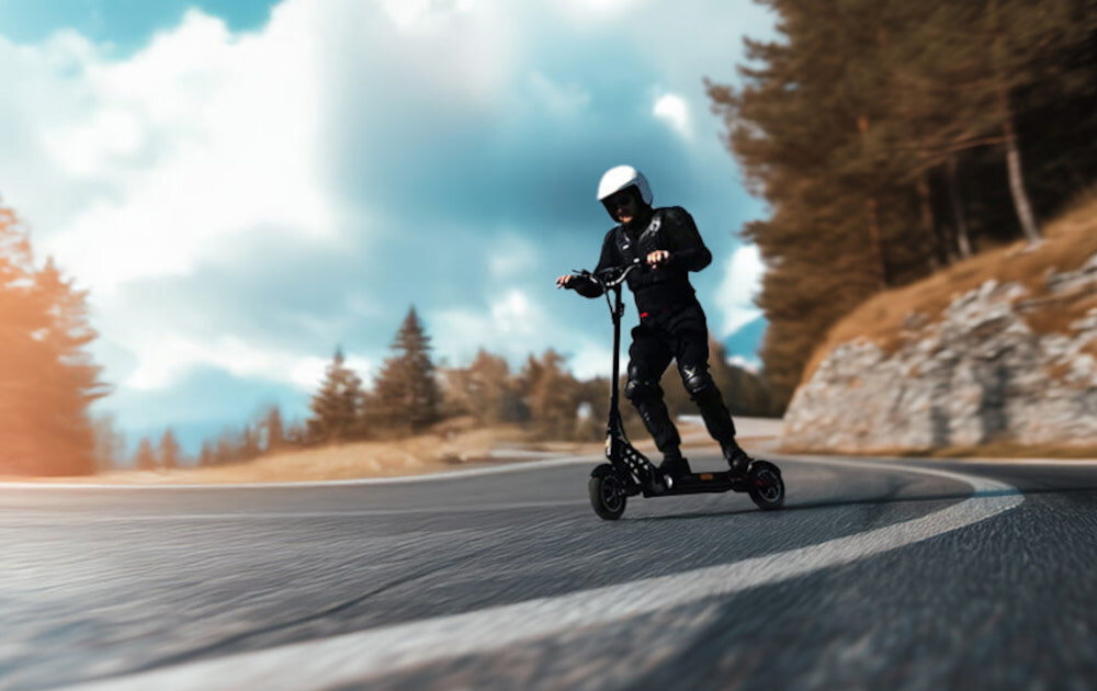 Hulajnoga elektryczna MOTUS Pro 10 Daytona 65km 2x1000W Amortyzacja 10 cali Czarny ulica po ulicy jedzie osoba w kasku na hulajnodze w tle drzewa skały dwa wydajne silniki o mocy 1000 W każdy cicha praca dynamiczna jazda moc szczytowa 1300 W pokonanie wzniesień o nachyleniu do 15% maksymalna prędkość do 20 km/h