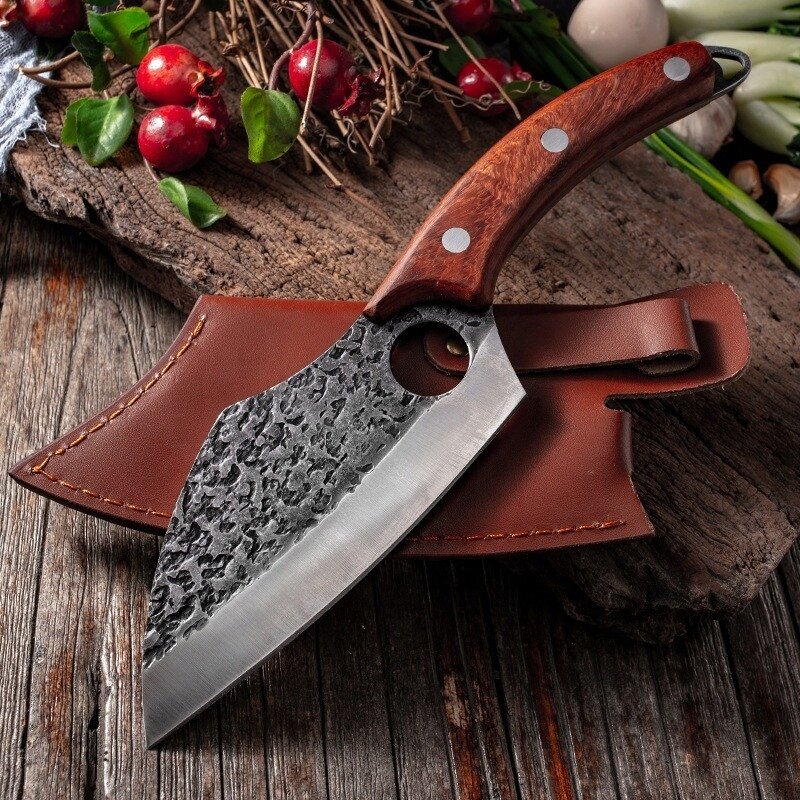 Na drewnianym stole leży nóż kuchenny z ciemnym, drewnianym uchwytem i ostrzem o młotkowanej fakturze, obok skórzanej pochwy. W tle widoczne są gałązki z czerwonymi owocami i zielonymi liśćmi.