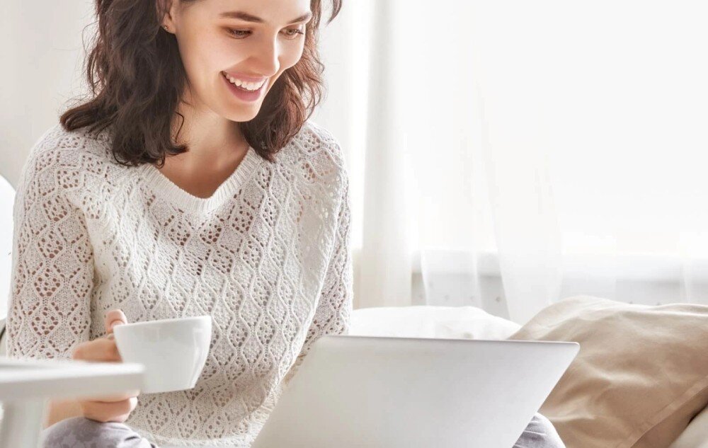 Router HUAWEI 4G Mobile Wi-Fi 5 E5586-326 Kobieta o ciemnych, kręconych włosach siedzi z uśmiechem przed otwartym laptopem. W jednej ręce trzyma biały kubek, a jej wzrok jest skierowany na ekran komputera. Scena jest jasna i utrzymana w domowej atmosferze