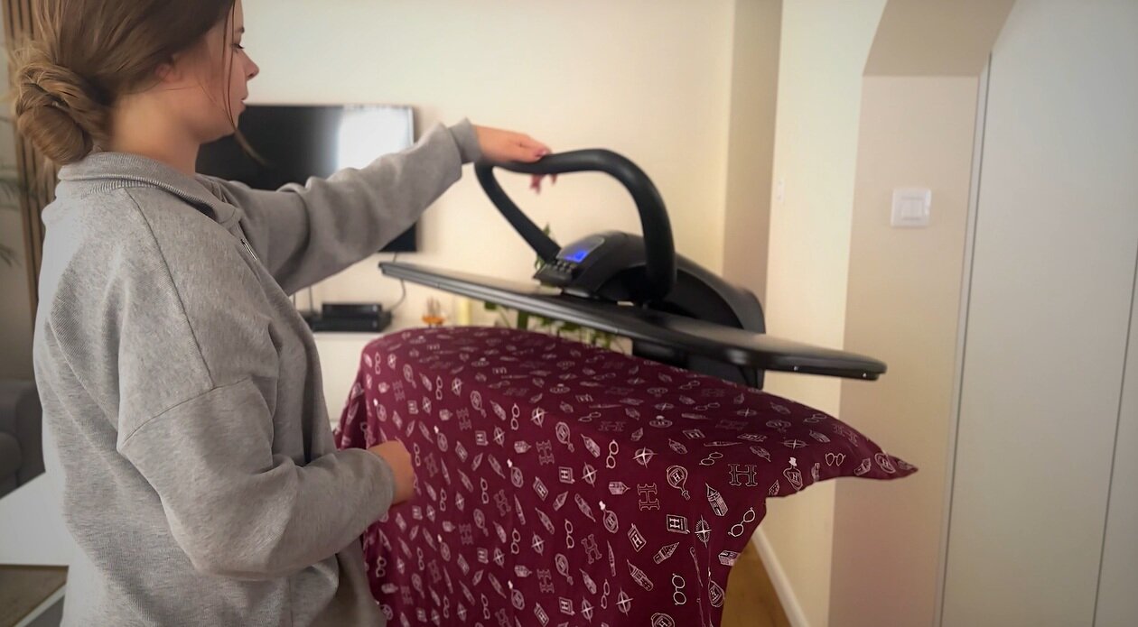 Osoba używa systemu do prasowania, trzymając uchwyt urządzenia nad bordowym materiałem z białymi wzorami. W tle widoczny jest telewizor i jasne ściany pomieszczenia.