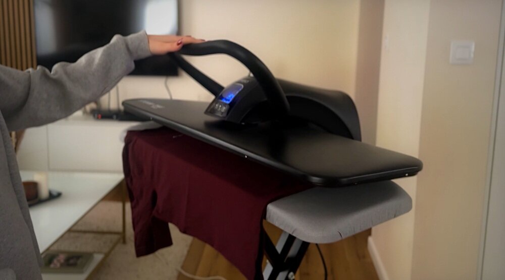 Osoba używa systemu do prasowania z czarną powierzchnią, na której znajduje się bordowa koszulka. W tle widoczny jest telewizor i część salonu.