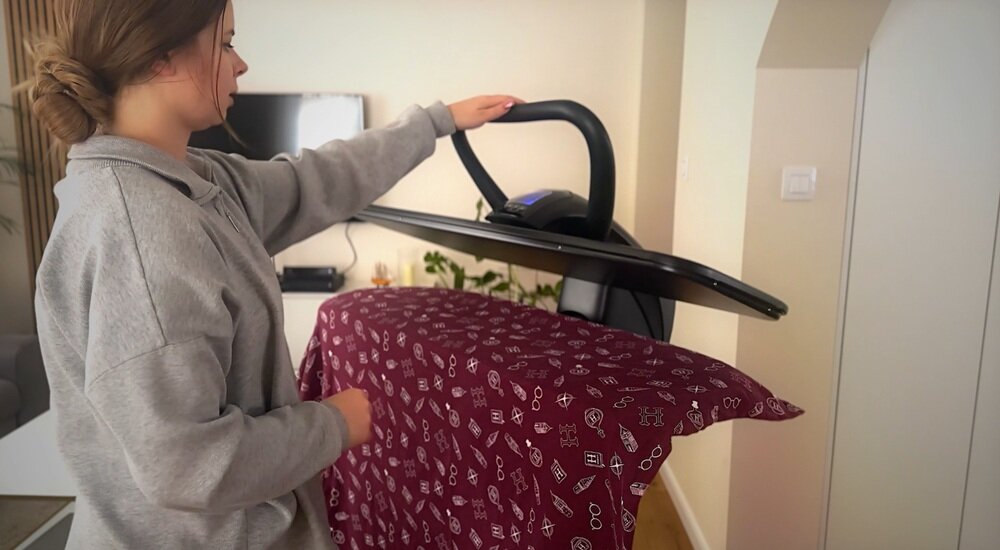 Osoba używa systemu do prasowania z czarną deską i uchwytem. Na desce znajduje się bordowy materiał z białymi wzorami.