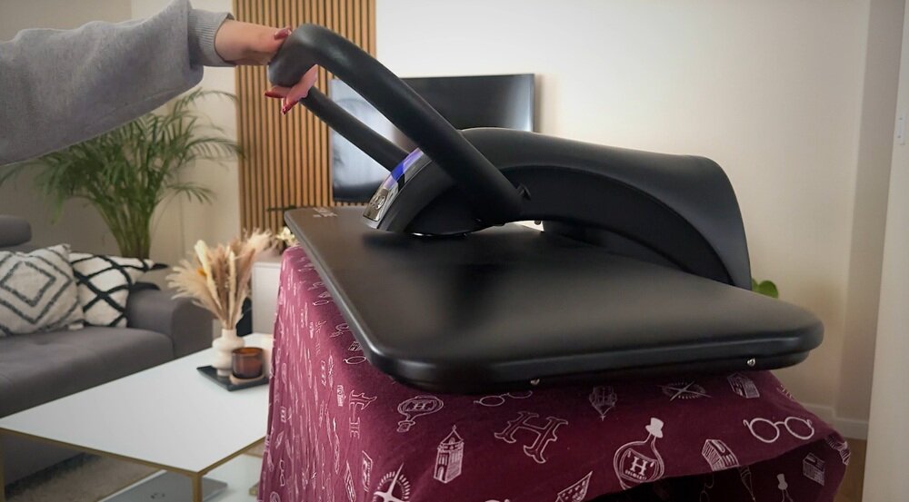 Na obrazie widać system do prasowania w kolorze czarnym, umieszczony na desce pokrytej bordowym materiałem z białymi wzorami. W tle znajduje się salon z rośliną, sofą i telewizorem.