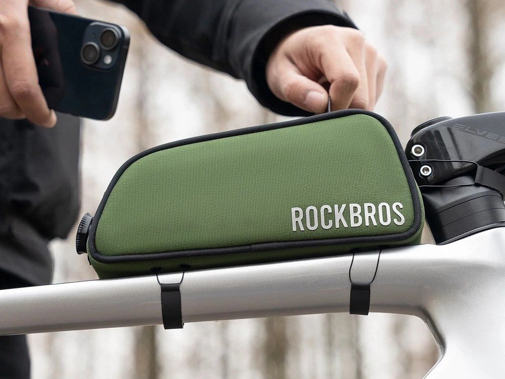 Zielona torba rowerowa marki ROCKBROS zamocowana na ramie roweru. Obok widoczna jest ręka trzymająca smartfon.