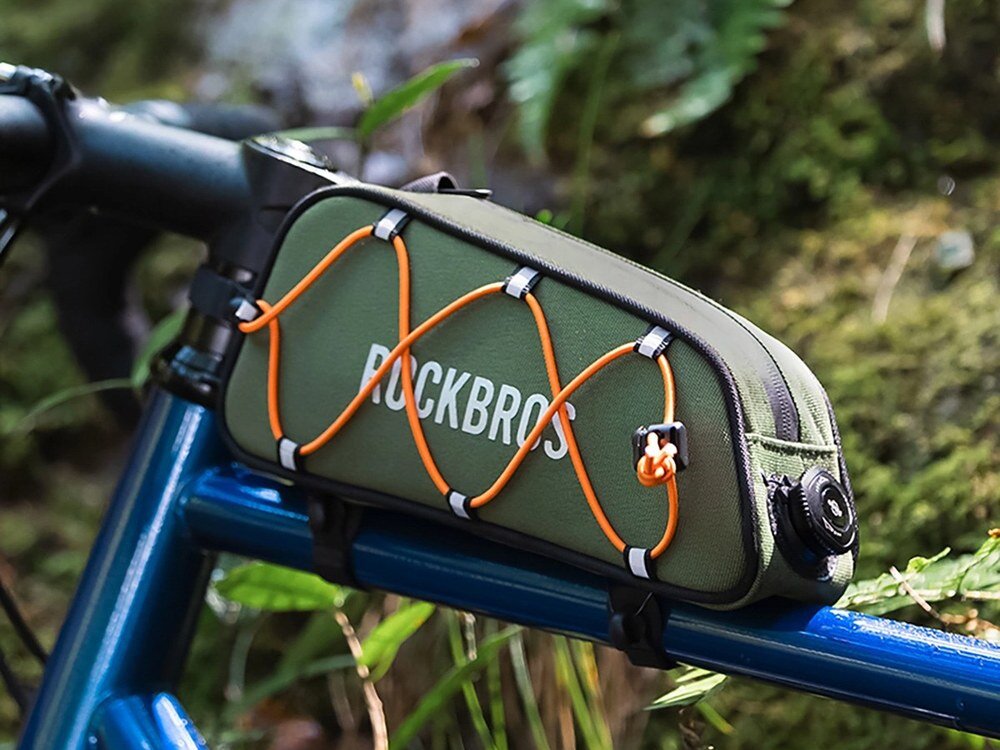 Zielona torba rowerowa zamontowana na ramie roweru z widocznym napisem \'ROCKBROS\' i pomarańczowymi sznurkami na wierzchu. W tle widoczne są liście i naturalne otoczenie.