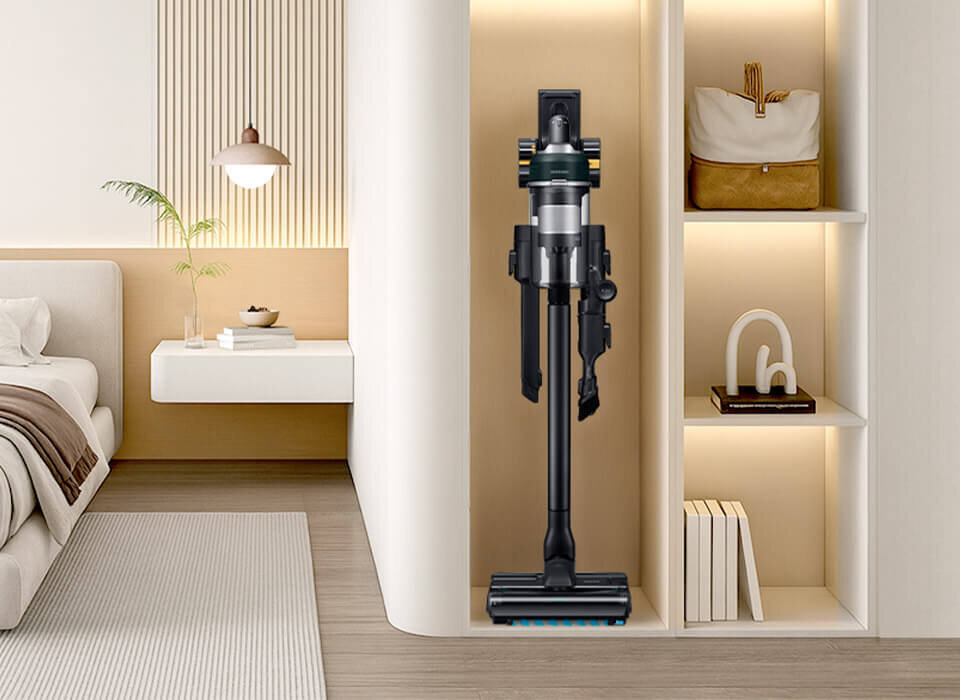 Wnętrze nowoczesnego pokoju z odkurzaczem pionowym umieszczonym w wnęce regału. Obok znajduje się łóżko, stolik z książkami i rośliną oraz półki z dekoracjami.