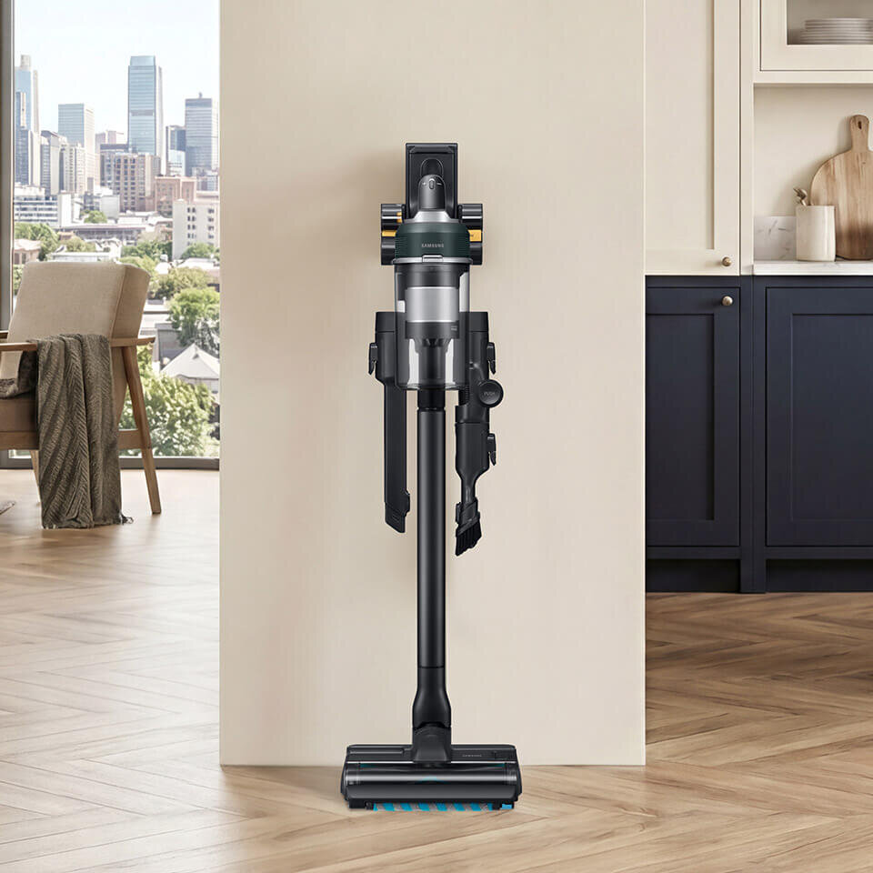 Na obrazie widoczny jest odkurzacz pionowy zamontowany na stacji dokującej przy ścianie w nowoczesnym wnętrzu. W tle widać duże okno z widokiem na miasto oraz część kuchni z szafkami i blatem.