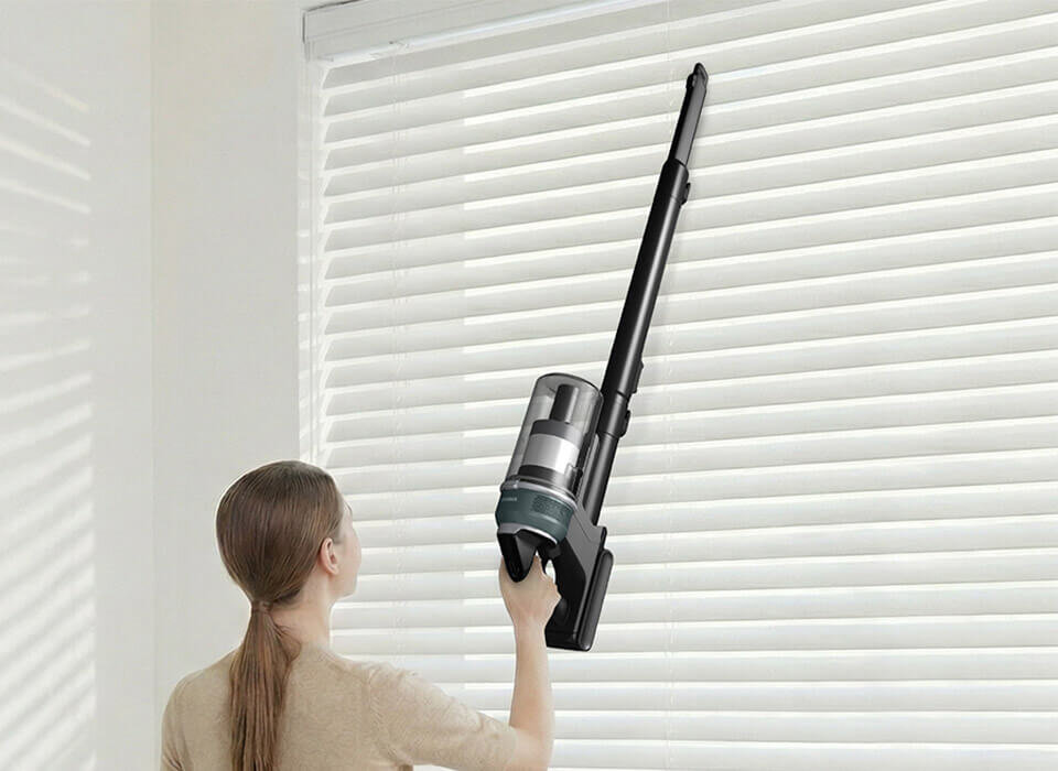 Osoba używa odkurzacza pionowego do czyszczenia żaluzji. Odkurzacz jest w kolorze czarnym i srebrnym.