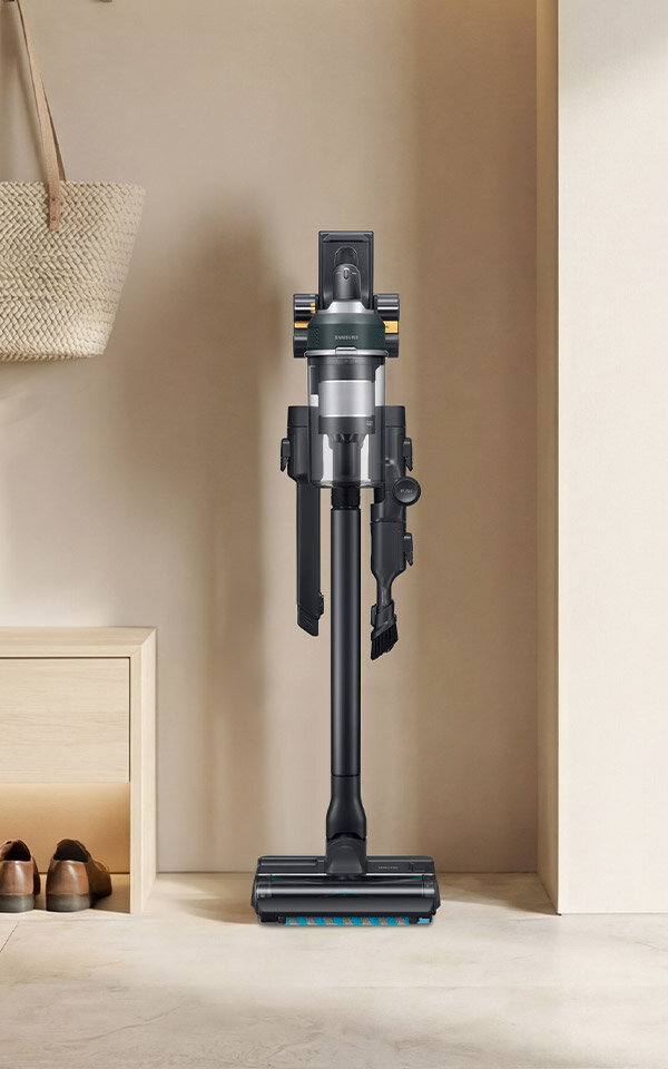 Na obrazie widoczny jest pionowo ustawiony odkurzacz bezprzewodowy w nowoczesnym wnętrzu. W tle znajduje się półka z parą butów i wiszącą torbą.