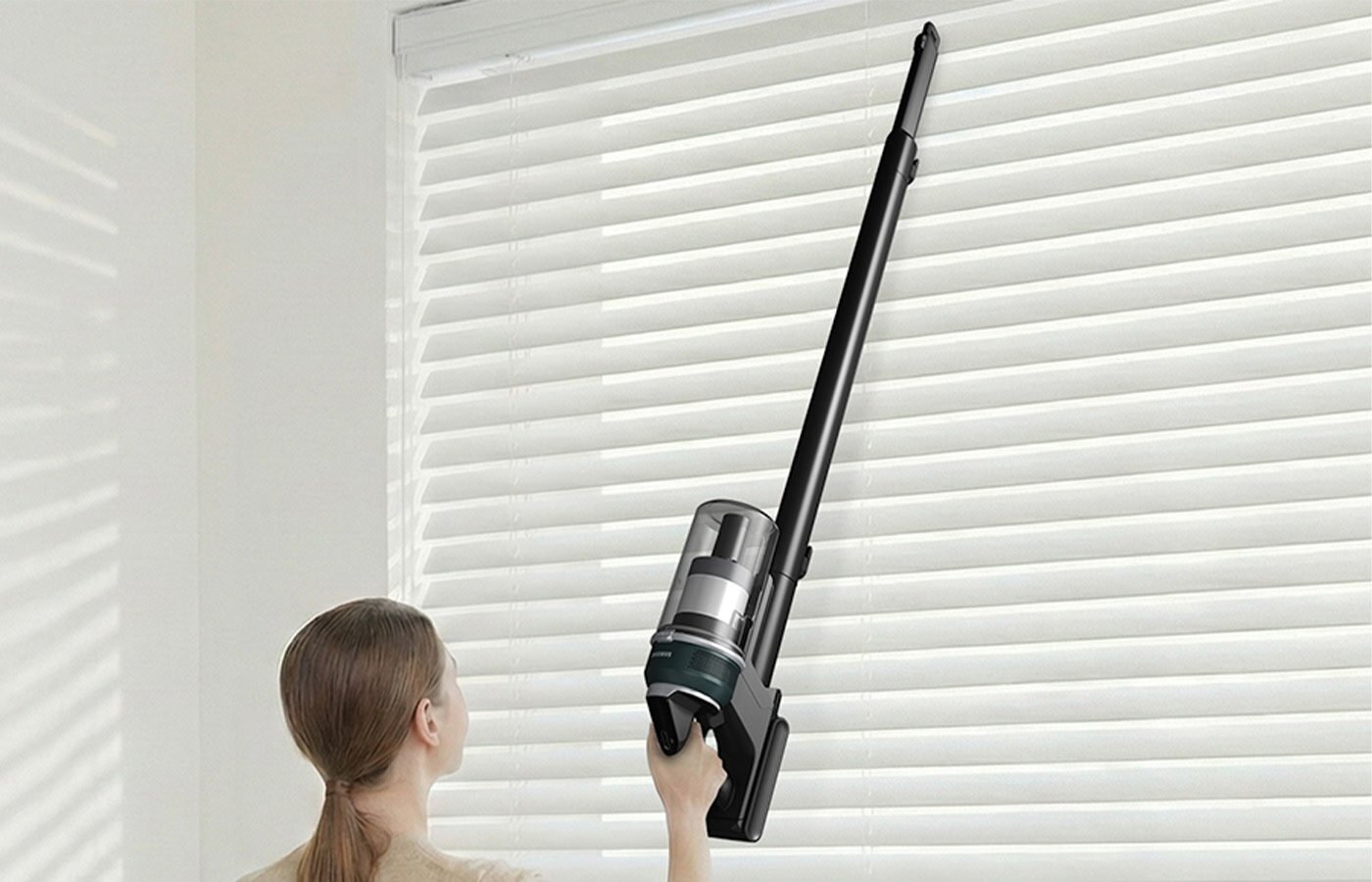 Osoba używa odkurzacza pionowego do czyszczenia żaluzji. Odkurzacz ma długą, czarną rurę i przezroczysty pojemnik na kurz.