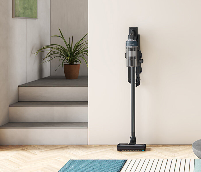 Na obrazie widoczny jest odkurzacz pionowy zamontowany na ścianie w nowoczesnym wnętrzu. Obok znajduje się roślina w doniczce na schodach.