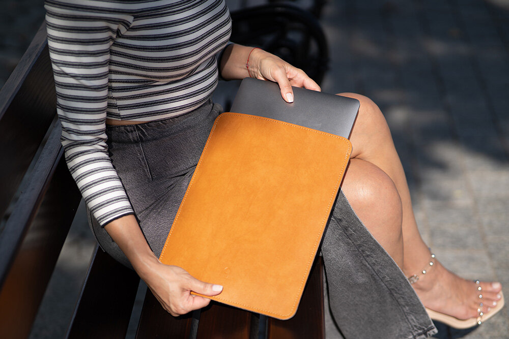 Osoba siedzi na ławce, trzymając jasnobrązowe etui na laptopa, z którego wystaje ciemnoszary laptop. Osoba ma na sobie bluzkę w paski i ciemne spodnie.