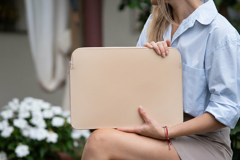 Osoba trzyma beżowe etui na laptopa, siedząc na zewnątrz. W tle widoczne są białe kwiaty i rozmyte elementy otoczenia.