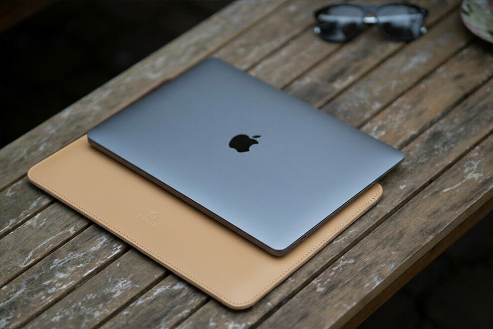 Na drewnianym stole leży srebrny laptop z logo jabłka, umieszczony na beżowym etui. Obok znajdują się okulary przeciwsłoneczne.