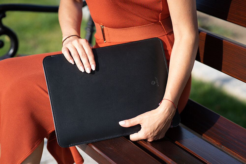 Osoba siedzi na ławce, trzymając czarne etui na laptopa. Etui ma gładką powierzchnię i widoczne logo producenta.