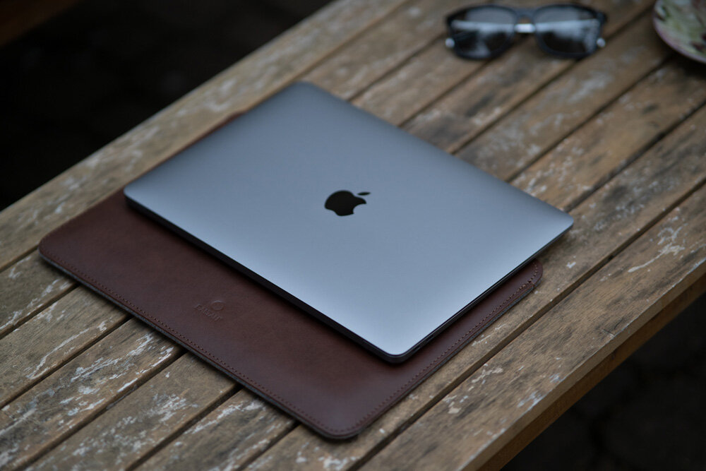 Na drewnianym stole leży srebrny laptop z logo jabłka, umieszczony na ciemnobrązowym etui. Obok znajdują się okulary przeciwsłoneczne.
