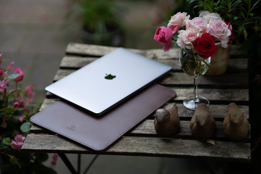Na drewnianym stole leży srebrny laptop z logo jabłka oraz ciemnobrązowe etui. Obok znajduje się szklanka z różowymi i czerwonymi różami oraz trzy figurki ptaków.