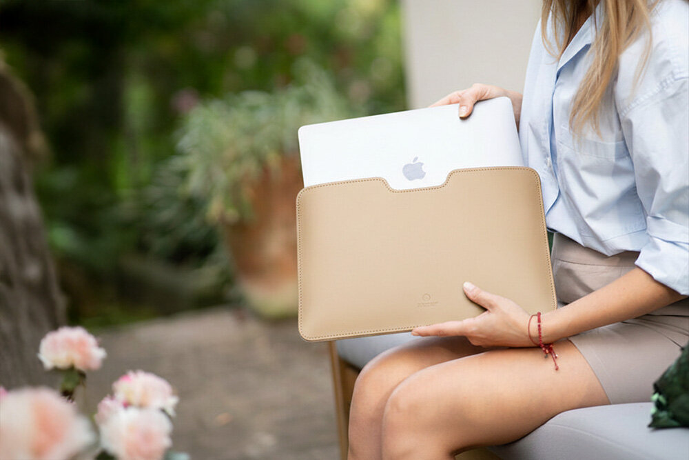 Osoba trzyma beżowe etui na laptopa, z którego wystaje srebrny laptop z logo jabłka. W tle widoczne są rozmyte rośliny i kwiaty.