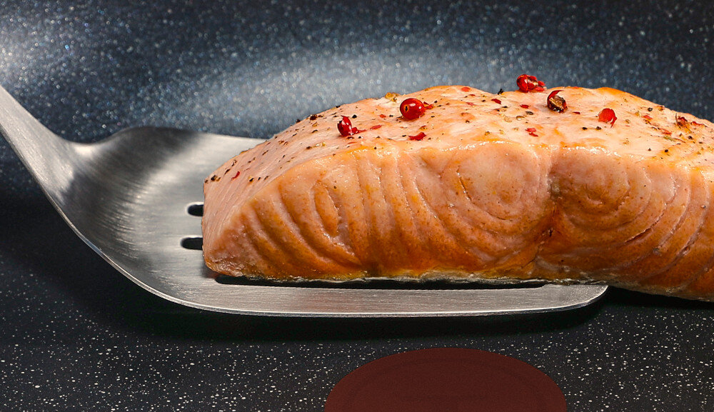 Na patelni widać kawałek smażonego łososia z przyprawami, leżący na metalowej łopatce. Powierzchnia patelni ma ciemny, nakrapiany wzór.